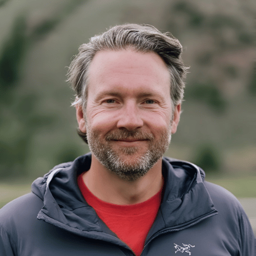 A man with grayish hair and beard, wearing a red shirt and dark jacket, stands outdoors with a blurred natural background.