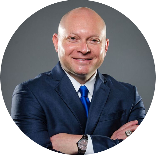 Bald man in a blue suit and tie stands with arms crossed, smiling, against a plain gray background.