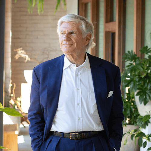 An older man with white hair wearing a blue suit and white shirt stands indoors near plants, looking to the side.