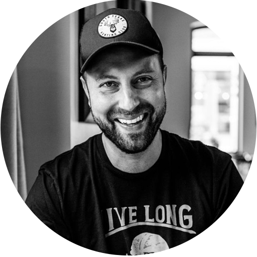 Smiling man in a baseball cap and t-shirt sits indoors, facing the camera. The photo is black and white.