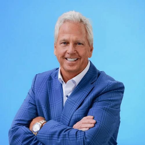 A man with white hair wearing a blue checked suit jacket and white shirt stands against a plain blue background with his arms crossed, smiling at the camera.