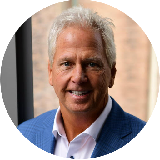 A middle-aged man with short gray hair is wearing a blue suit jacket and white shirt, smiling at the camera with a blurred indoor background.